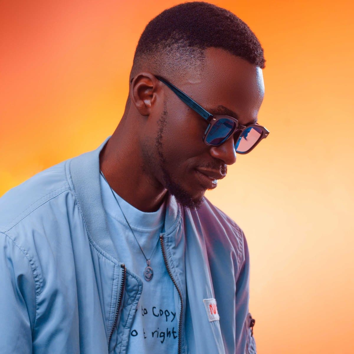 Ryan Synth in sunglasses and a light denim jacket against a warm orange studio backdrop